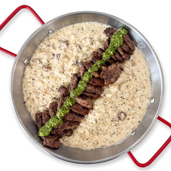 Grilled beef strips topped with green sauce over creamy mushroom rice in a metal skillet with red handles.