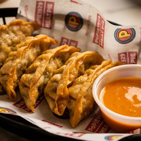 A basket of golden-brown fried dumplings with a side of orange dipping sauce on branded Popie1929 paper.