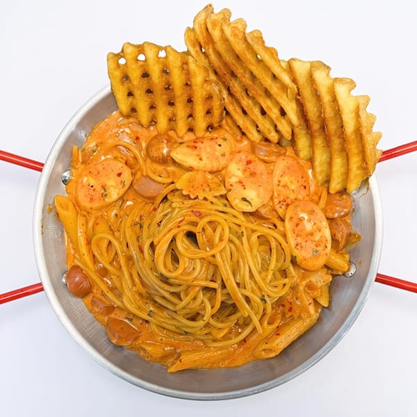 Overhead view of creamy orange pasta with sliced sausage, ravioli, tomatoes, and waffle fries in a metal pan.