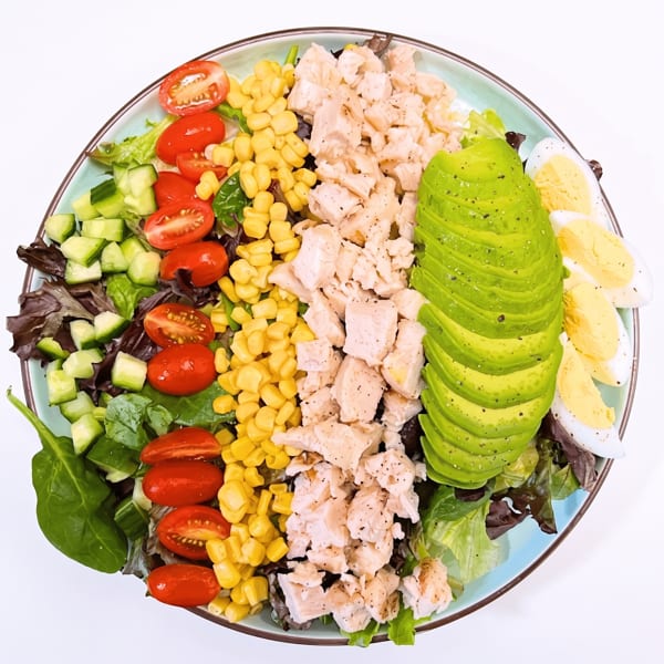 Colorful chicken salad with mixed greens, cucumber, tomatoes, corn, avocado slices, and hard-boiled egg.